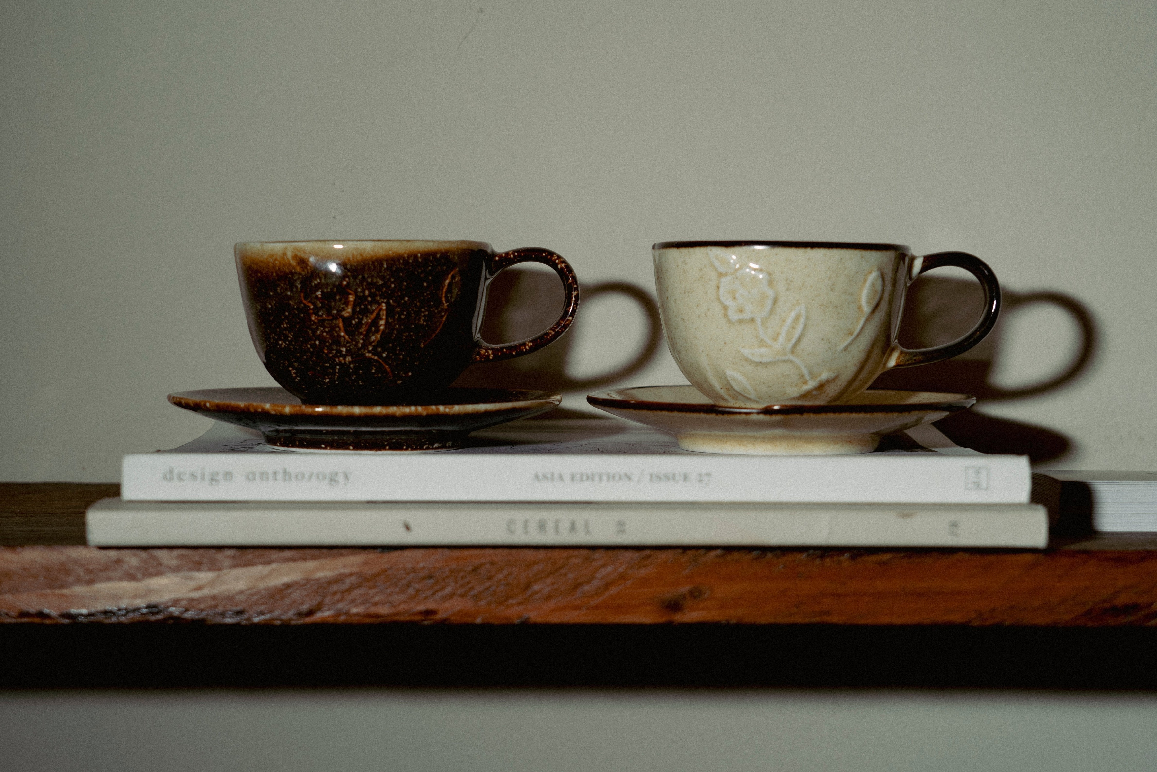 Our two Hana Kasumi ceramic cups in beige and brown, placed on top of a pile of magazines on a wooden shelf. 