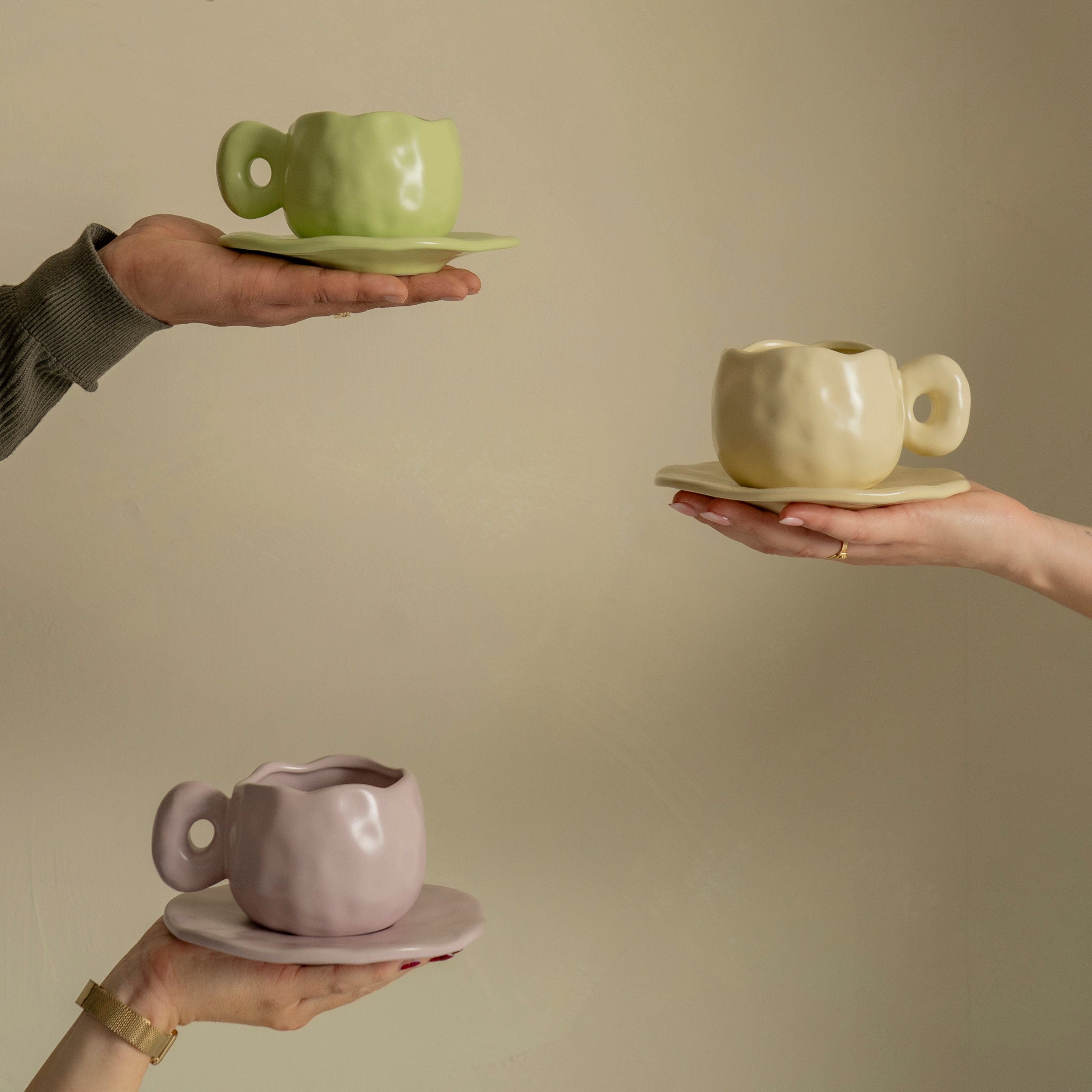 Picture of three hands, each holding a pastel color of our ceramic Curve mug: green, beige and purple. 